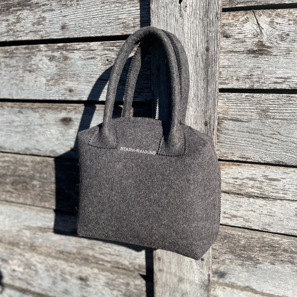 A gray felt handbag hangs on a wooden fence in sunlight.