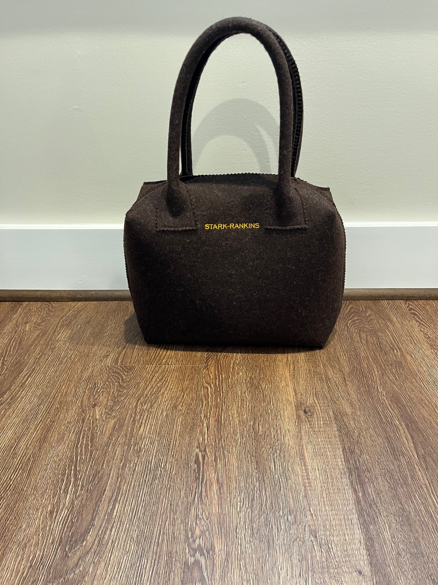 A dark brown handbag with "STARK-RANKINS" in gold lettering is placed on a wooden floor against a white wall.