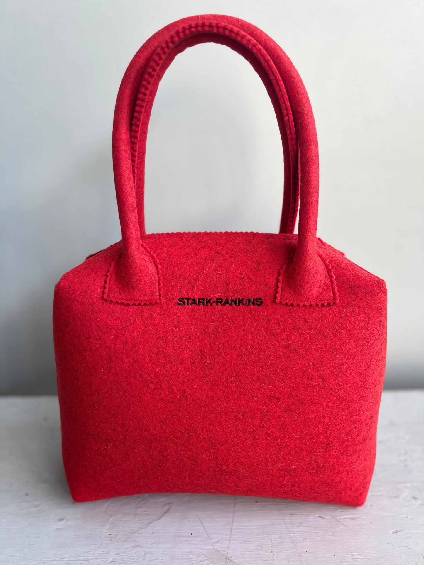 A red felt handbag with two handles and the words "STARK-RANKINS" on the front, placed against a plain background.
