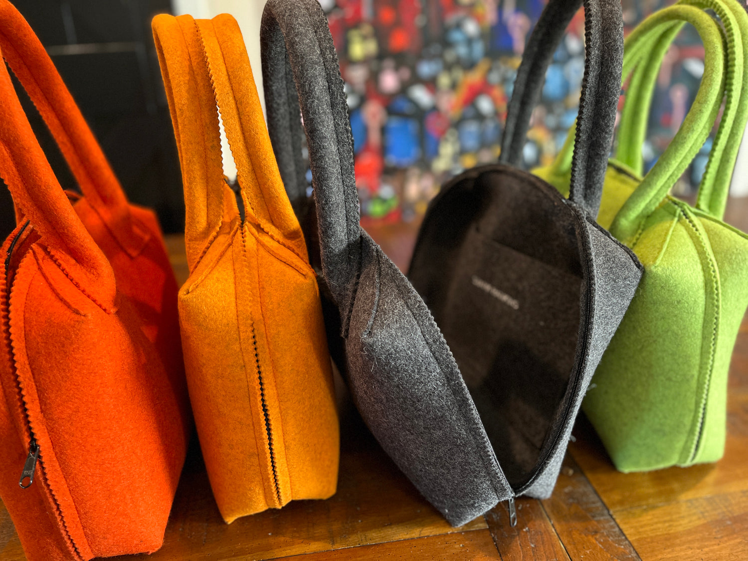 Four felt tote bags in orange, gray, and green are lined up on a wooden surface against a colorful, blurred background.