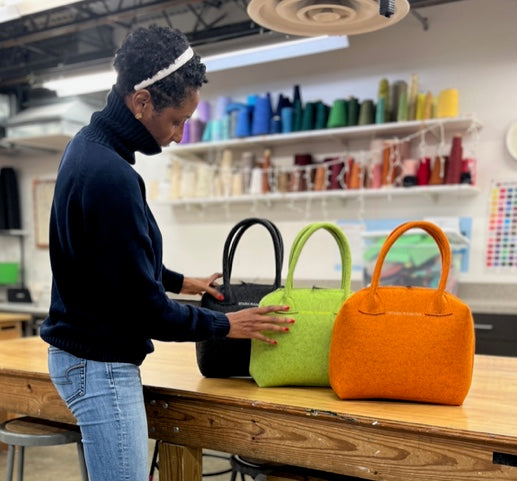 A person examines three handbags in black, green, and orange on a wooden table in a fabric workshop.