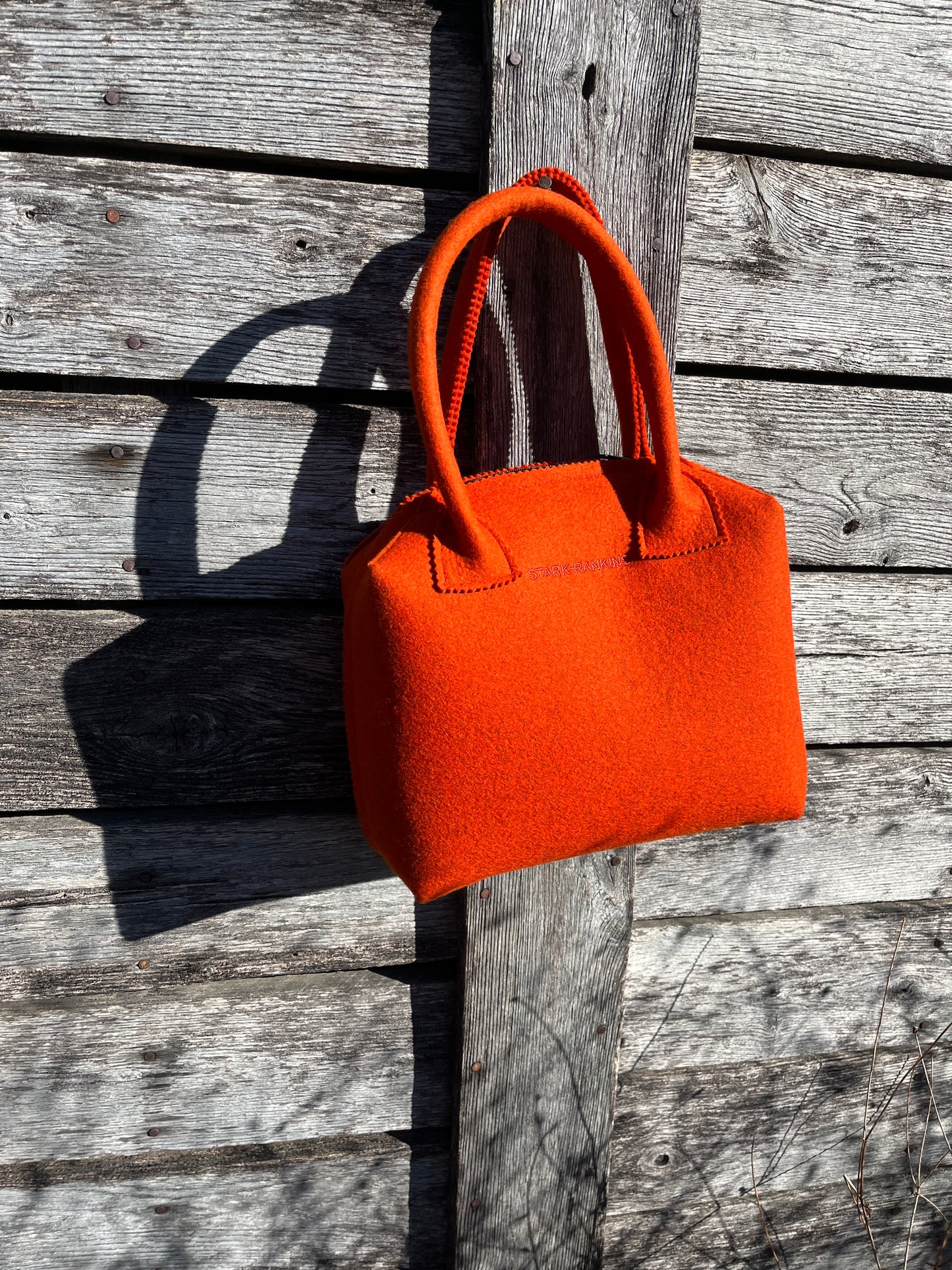 An orange felt handbag with two handles hangs on a weathered wooden wall in sunlight.
