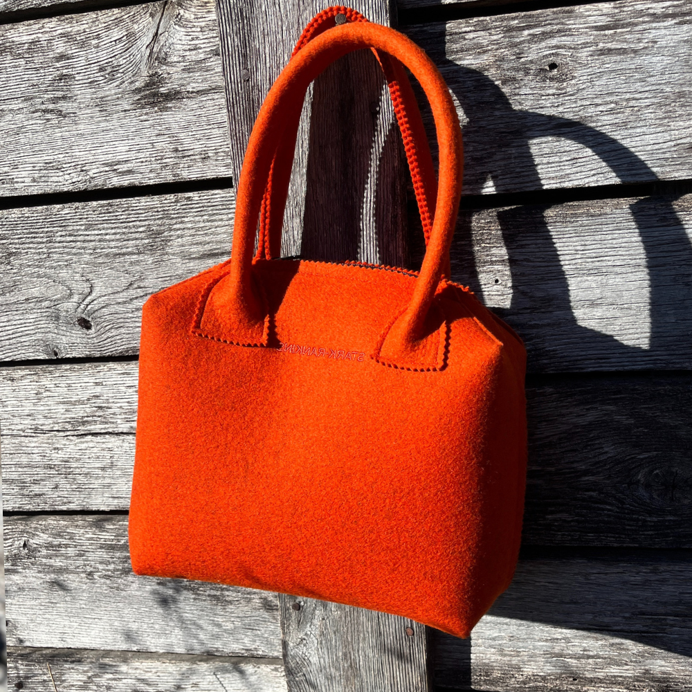 An orange felt handbag with two handles hangs against a weathered wooden wall, casting a shadow.