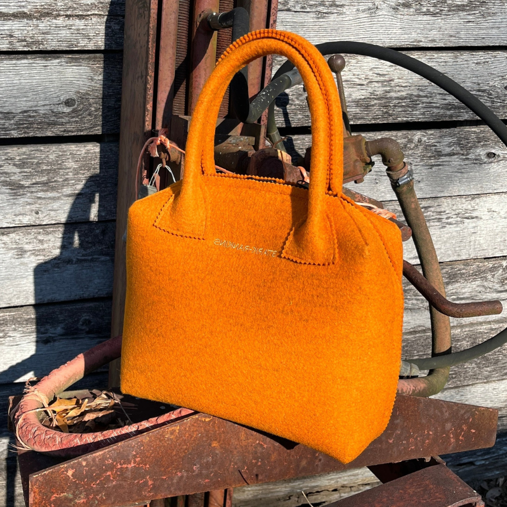 A bright orange handbag rests on a rusty metal surface, with a wooden wall and coiled rope in the background.