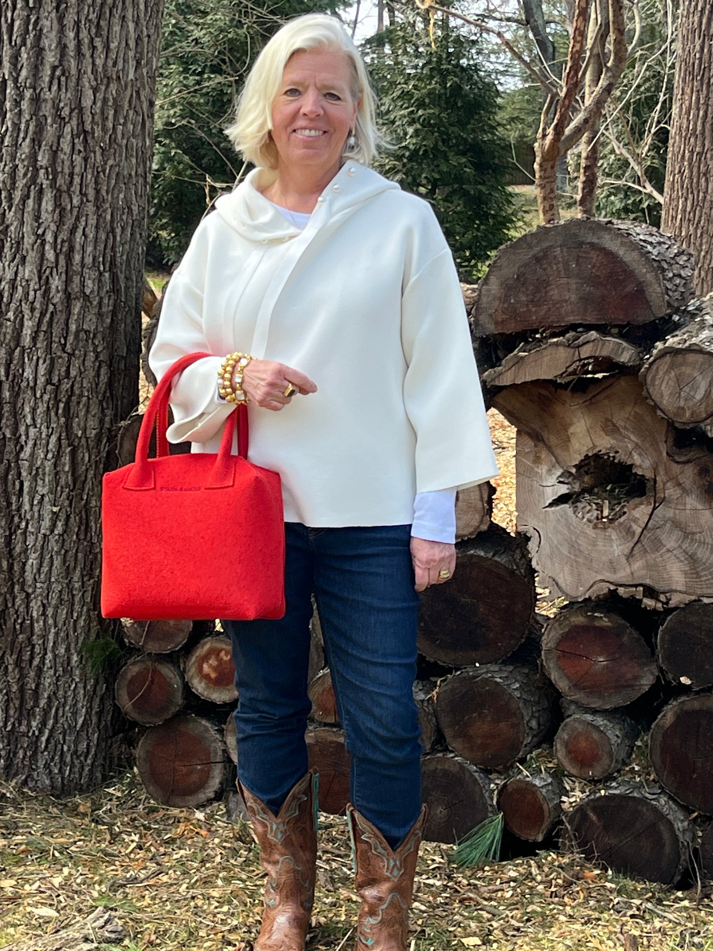 Person wearing a white hoodie and jeans stands by stacked logs, holding a red bag.