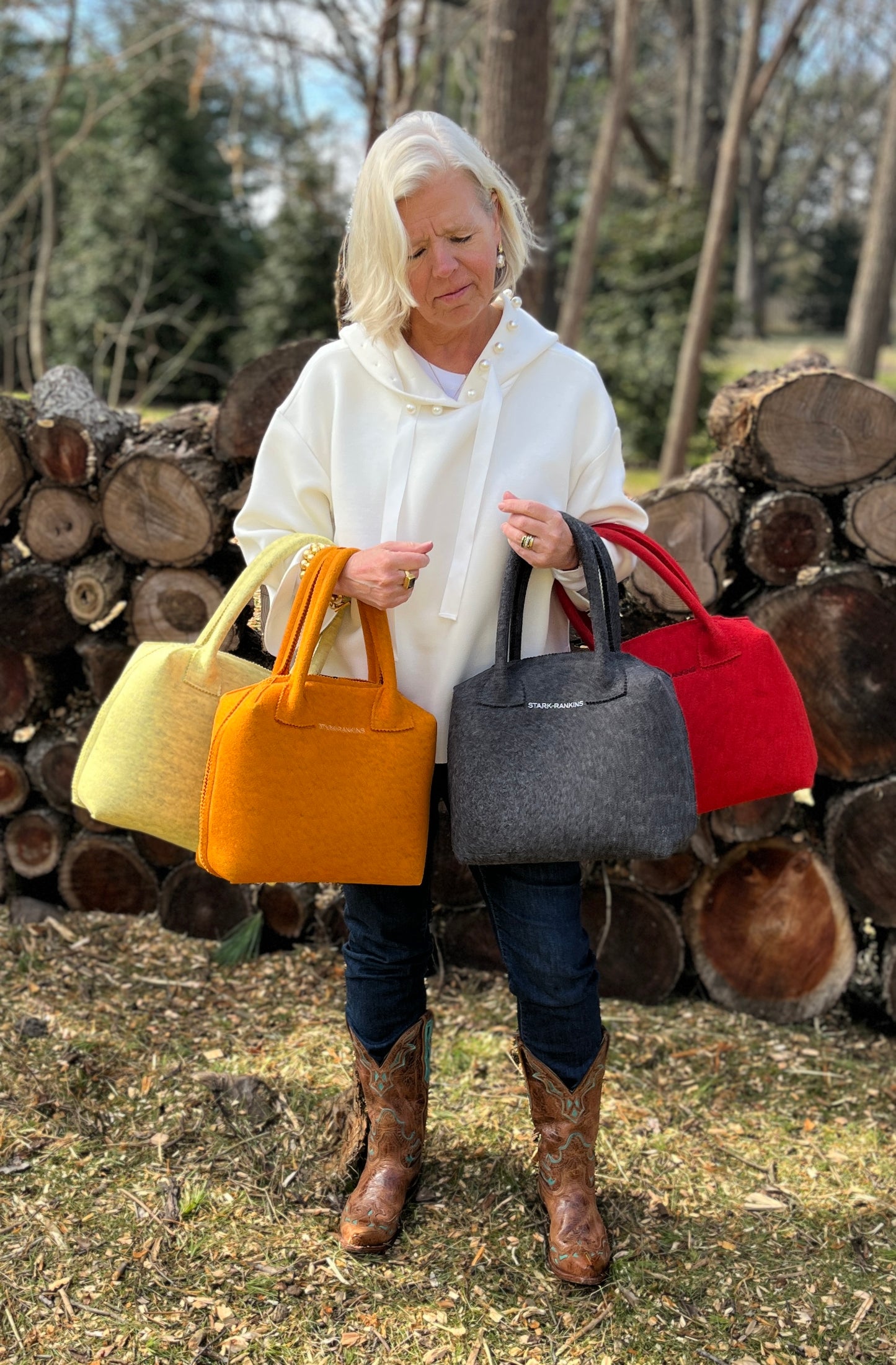 A person stands outdoors holding four colorful handbags in front of stacked logs.