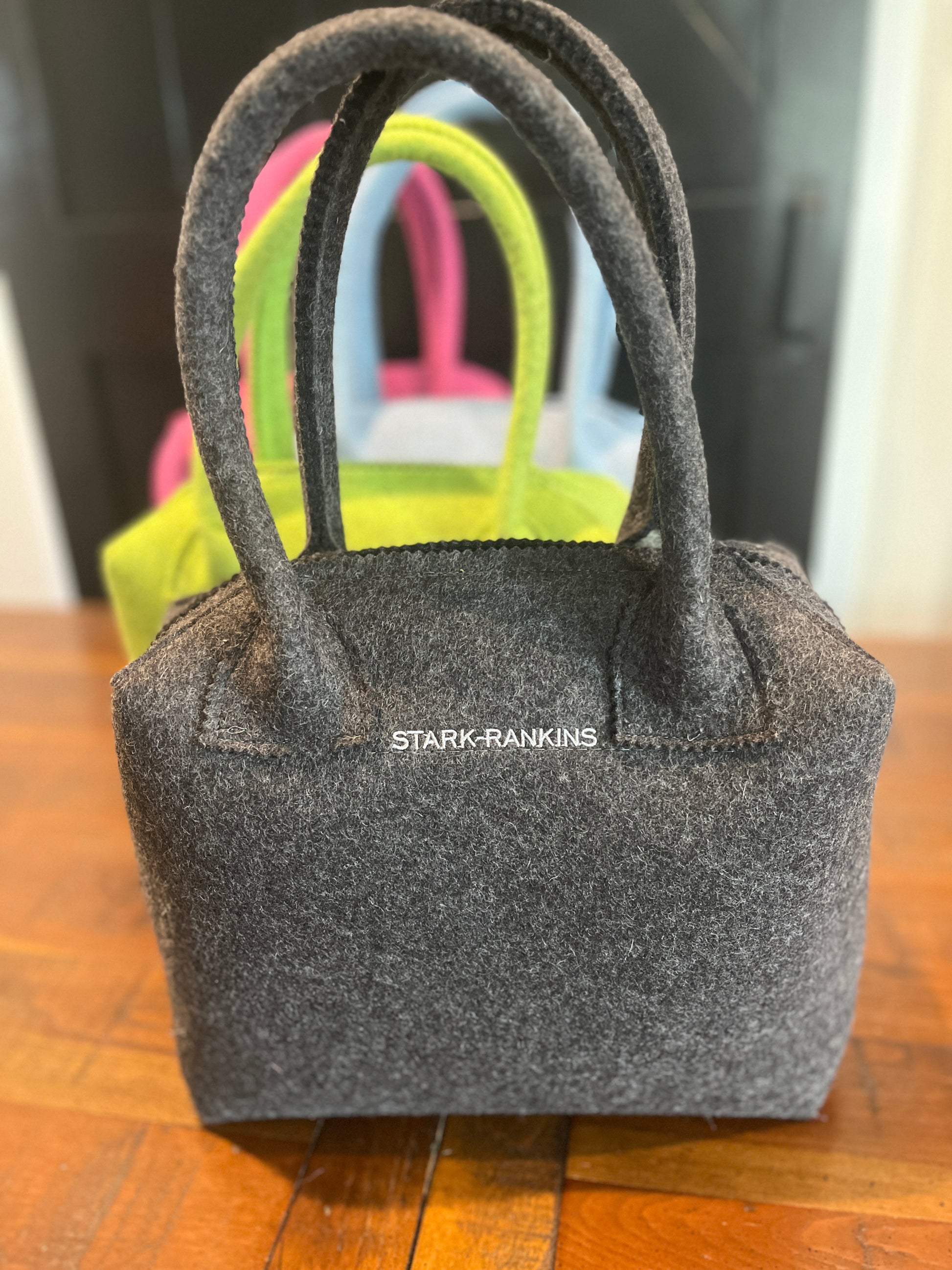 A gray felt handbag with "STARK-RANKINS" text is in focus, with a green and pink handbag blurred in the background. They are on a wooden surface.