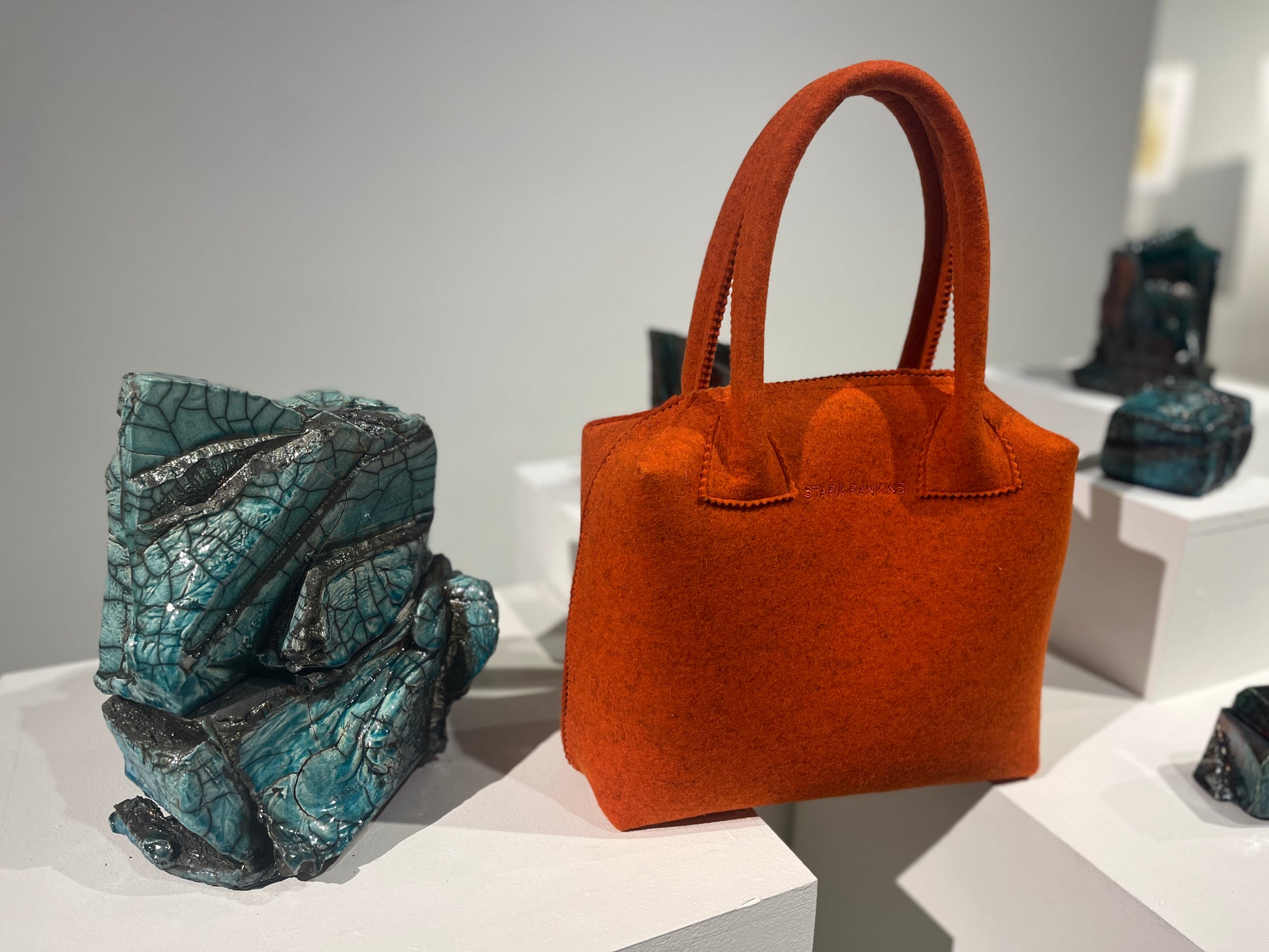 A vibrant orange handbag is displayed on a pedestal next to abstract turquoise sculptures in an exhibition setting.