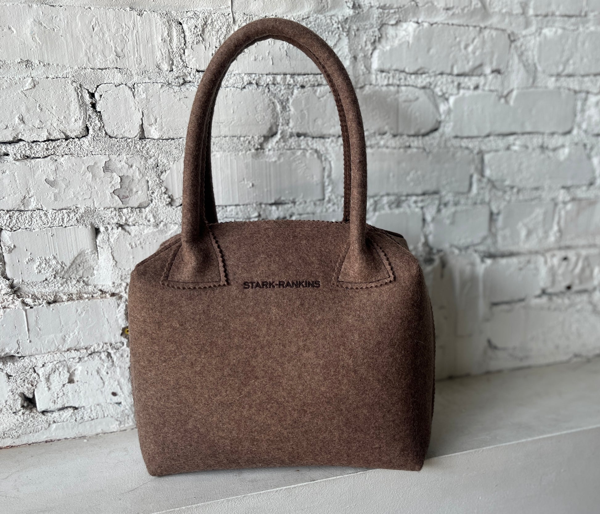 A brown felt handbag with two handles rests against a white brick wall.