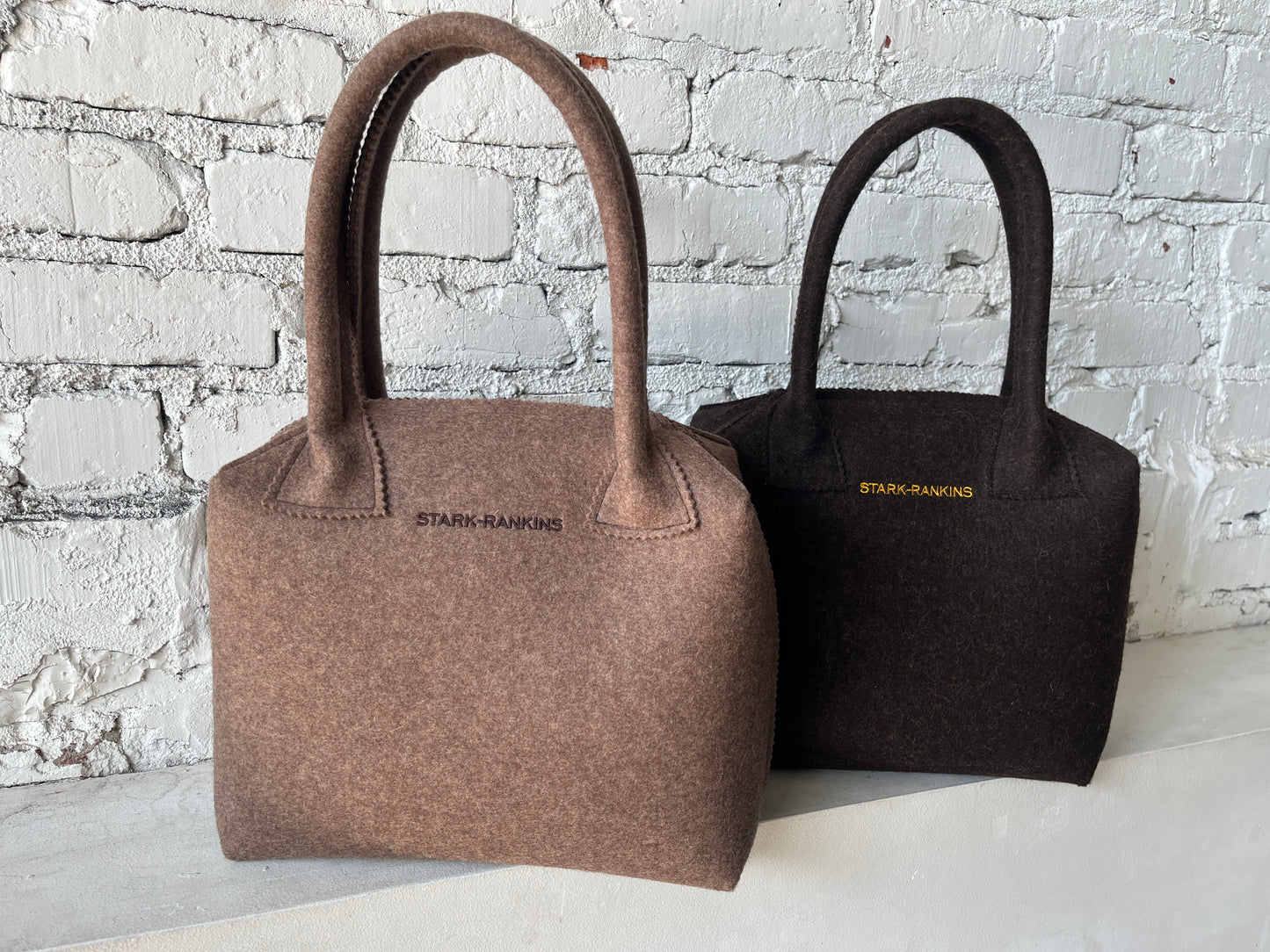 Two felt handbags on a white surface against a brick wall. One is brown, and the other is black, each with the text "STARK RANKINS" on the front.