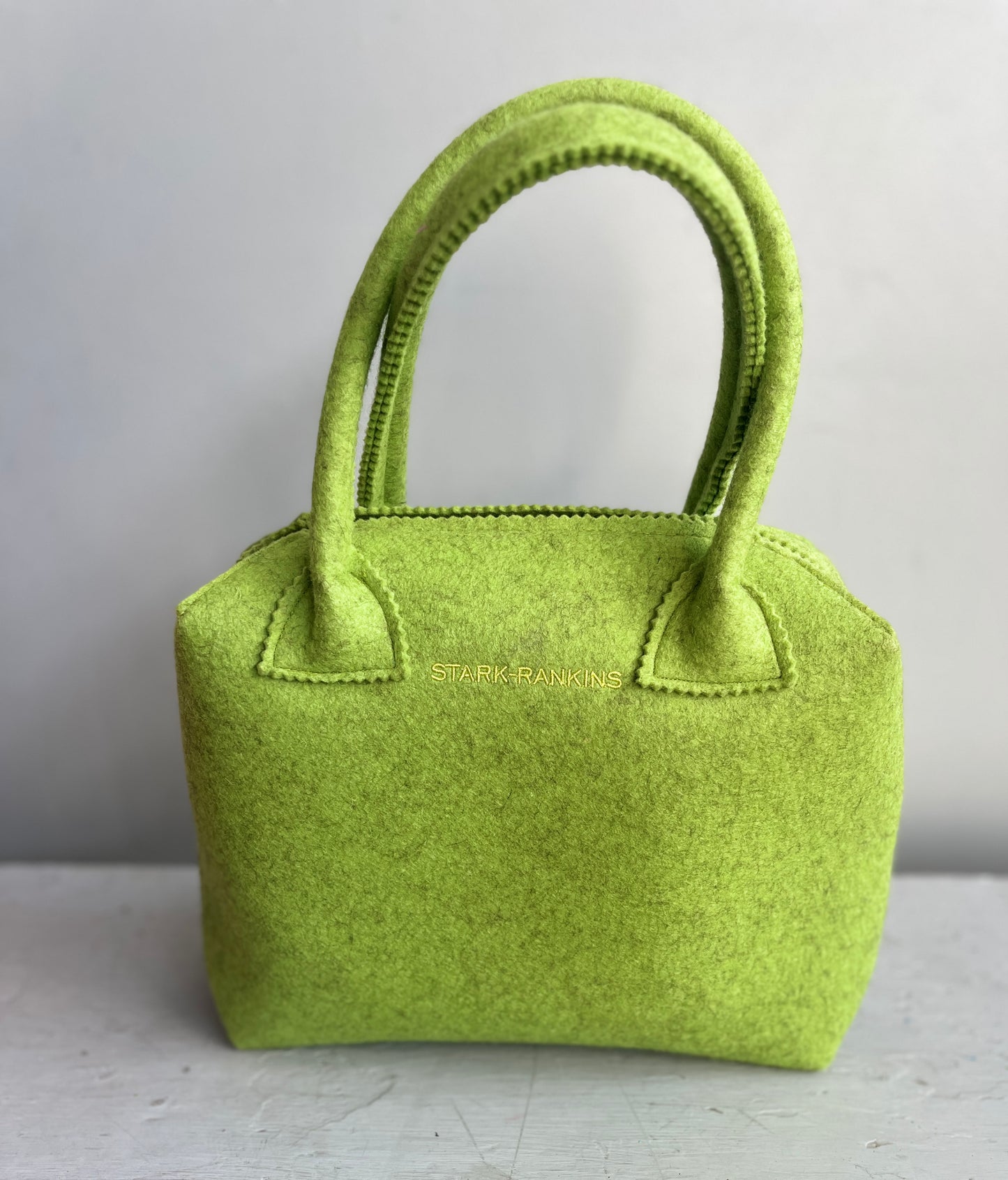 A small, lime green felt handbag with two handles. It rests against a plain background.