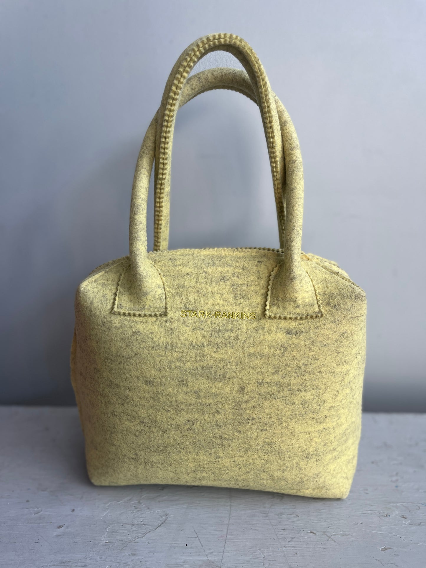 A yellow felt handbag with two handles placed on a gray surface against a gray background.