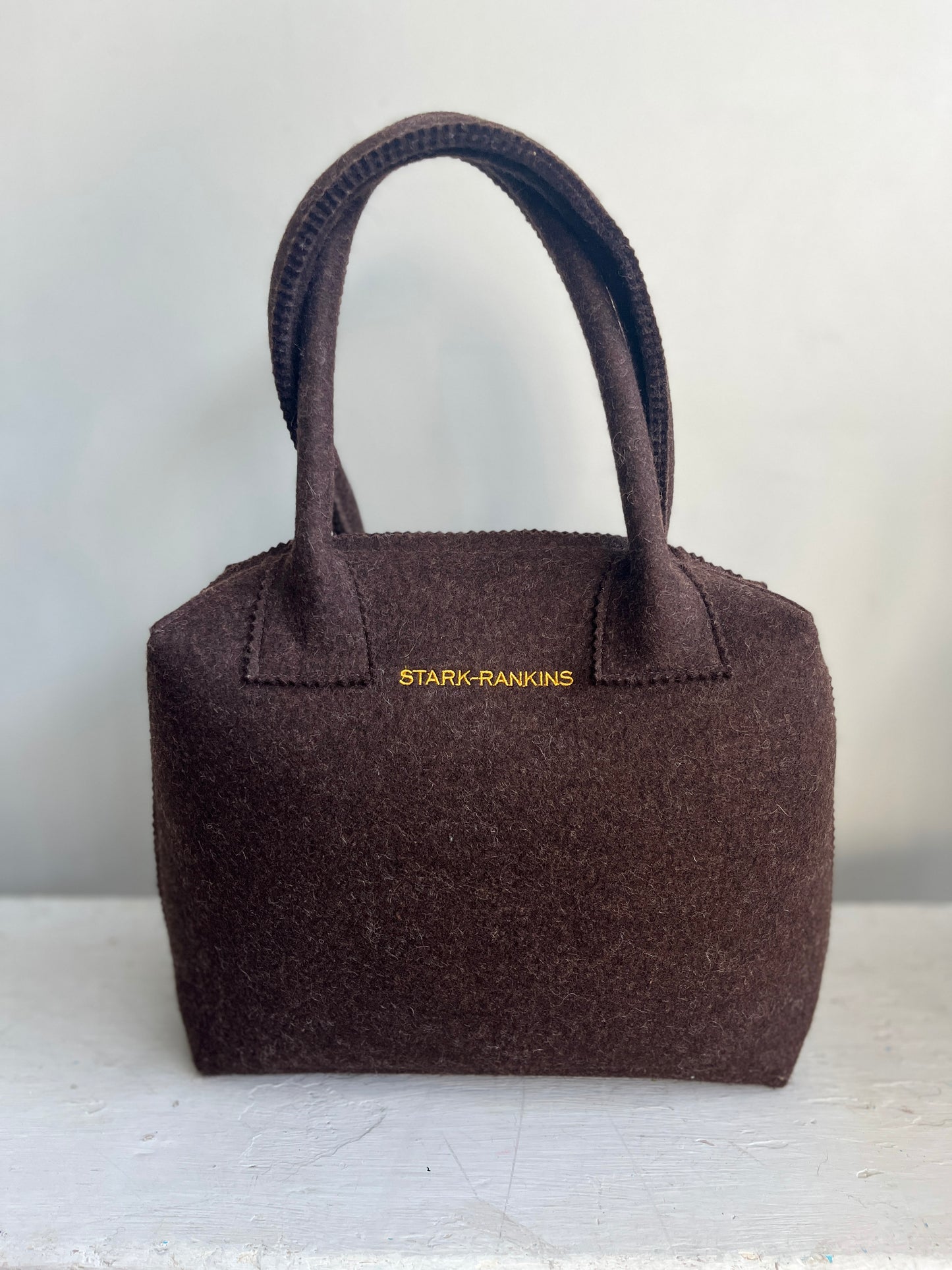 Brown felt handbag with two handles resting on a white surface, featuring the text "STARK-RANKINS" in yellow near the top.