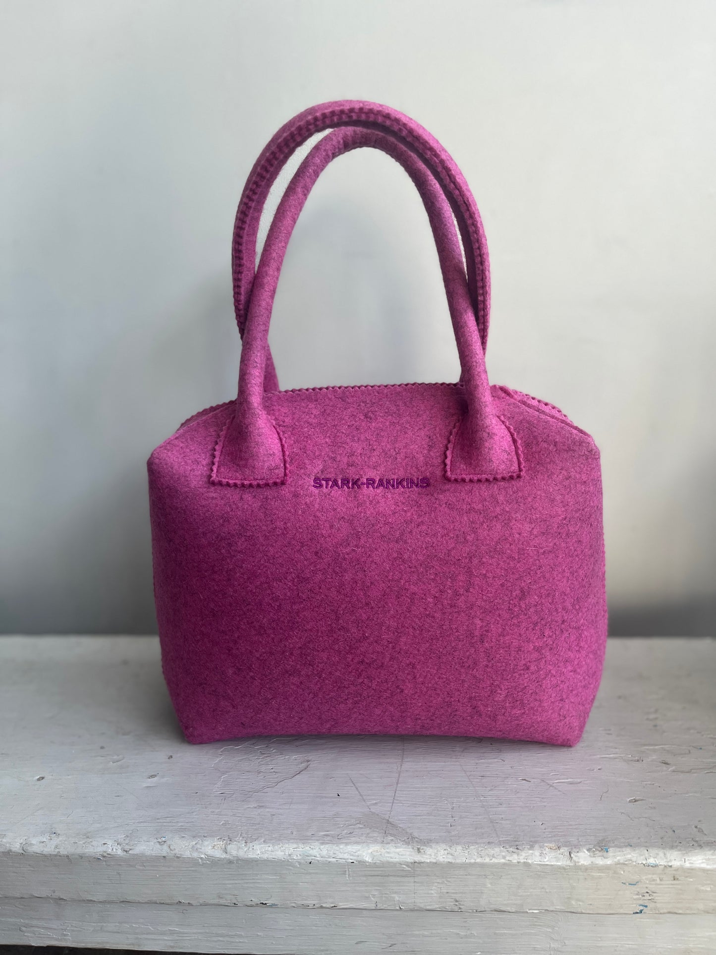 A pink felt handbag with two handles sits on a white surface. It has "Stark-Rankins" printed in small letters on the front.