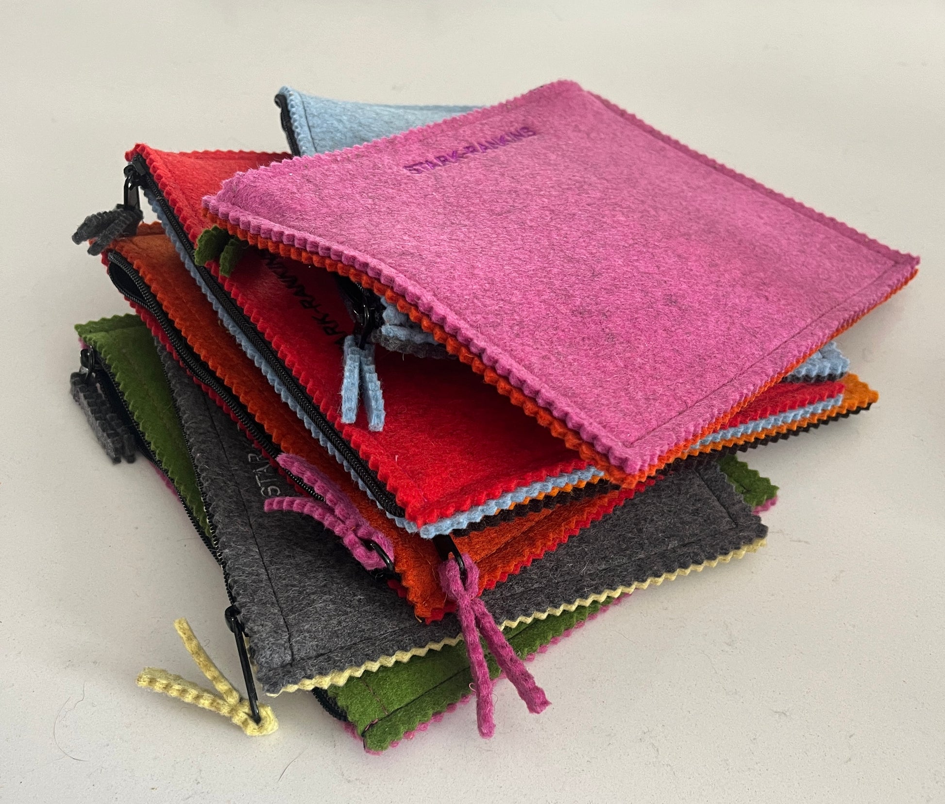 A stack of colorful felt zippered pouches, in shades of pink, red, orange, green, and gray, on a light surface.