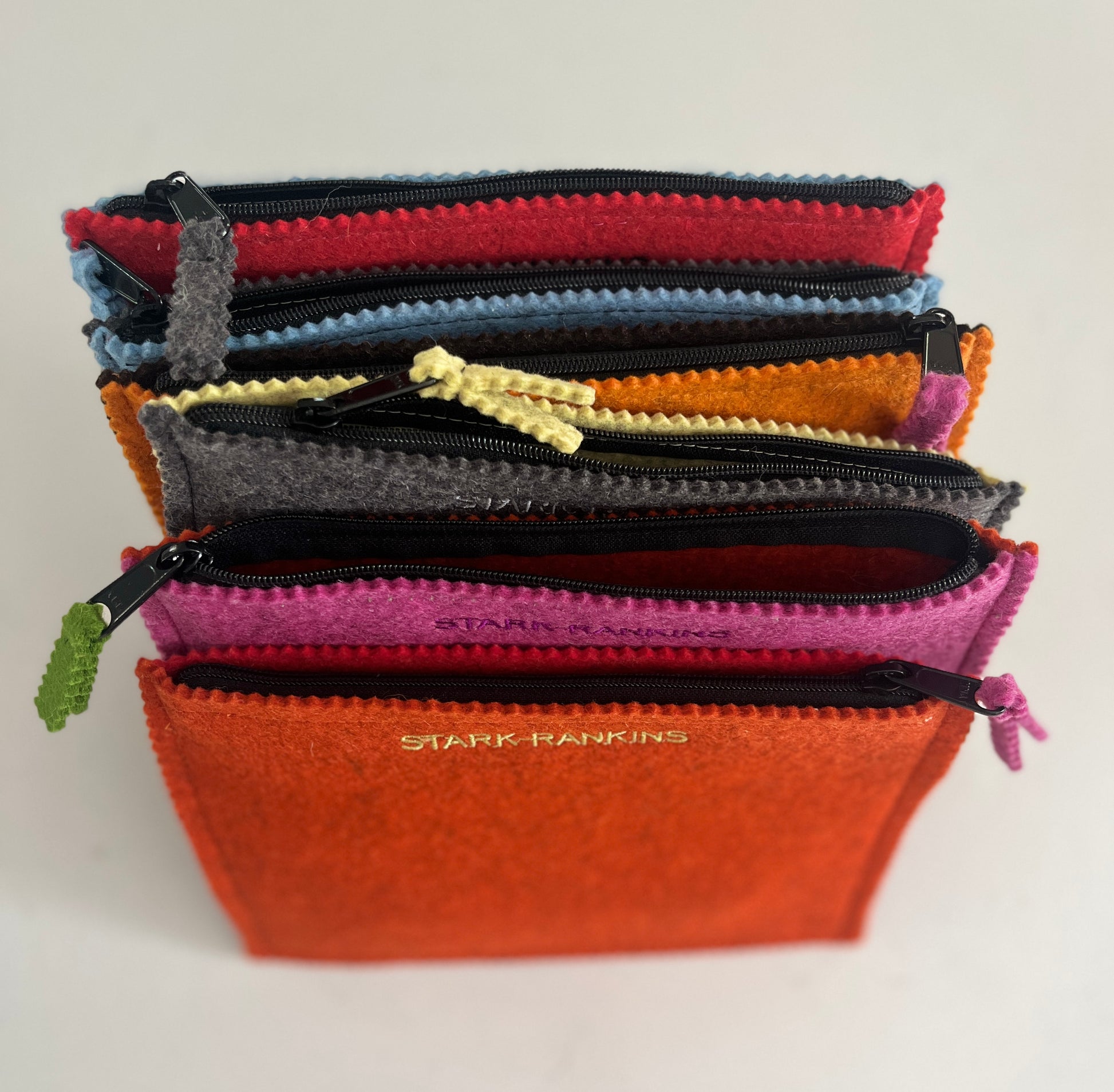 Stack of colorful zippered pouches arranged vertically, featuring various shades and textures, with a visible label reading "Stark Rankins" on the front orange pouch.