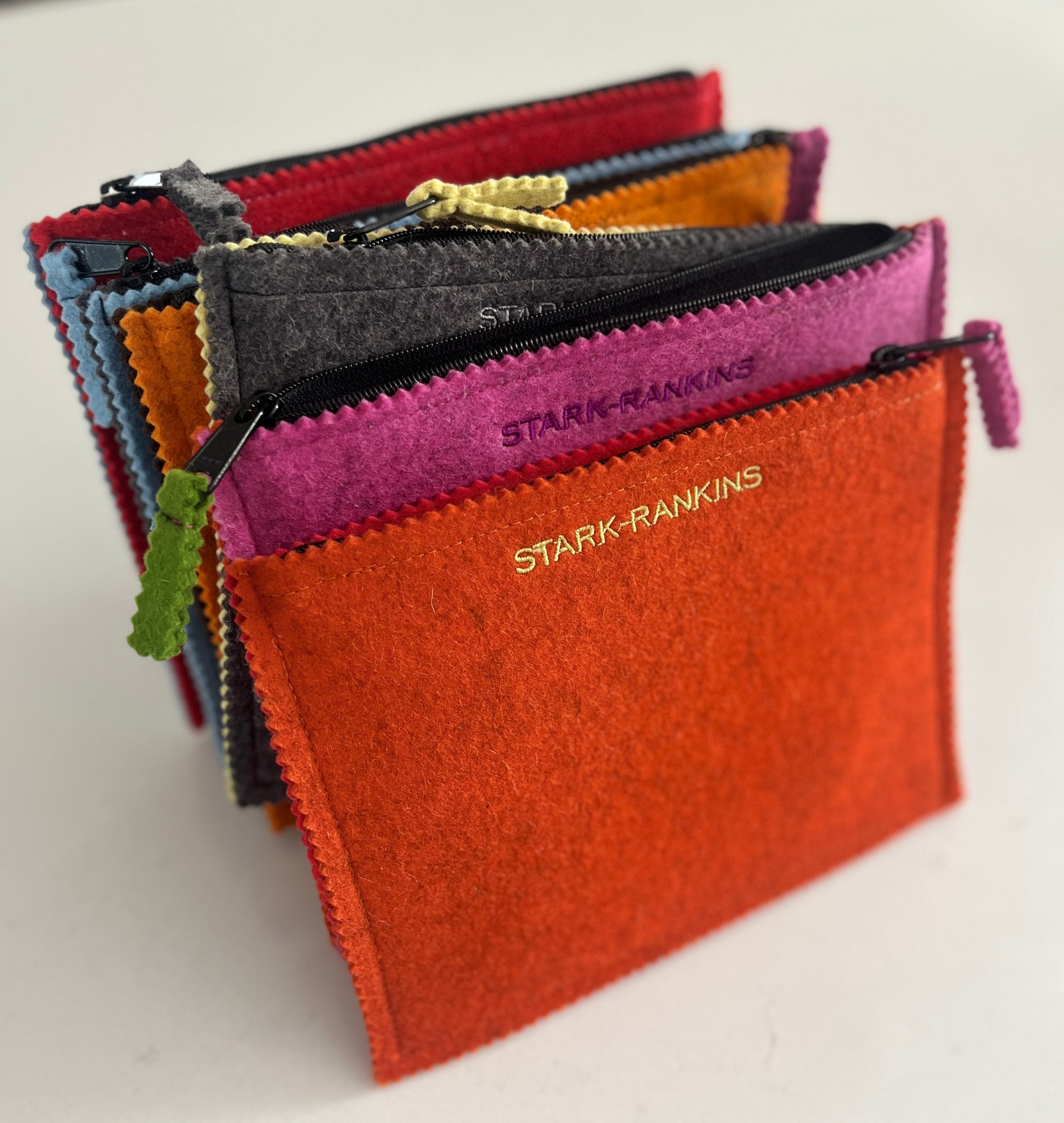 A collection of colorful felt pouches with zippers, stacked vertically. The front pouch is orange and has "STARK-RANKINS" embroidered on it.