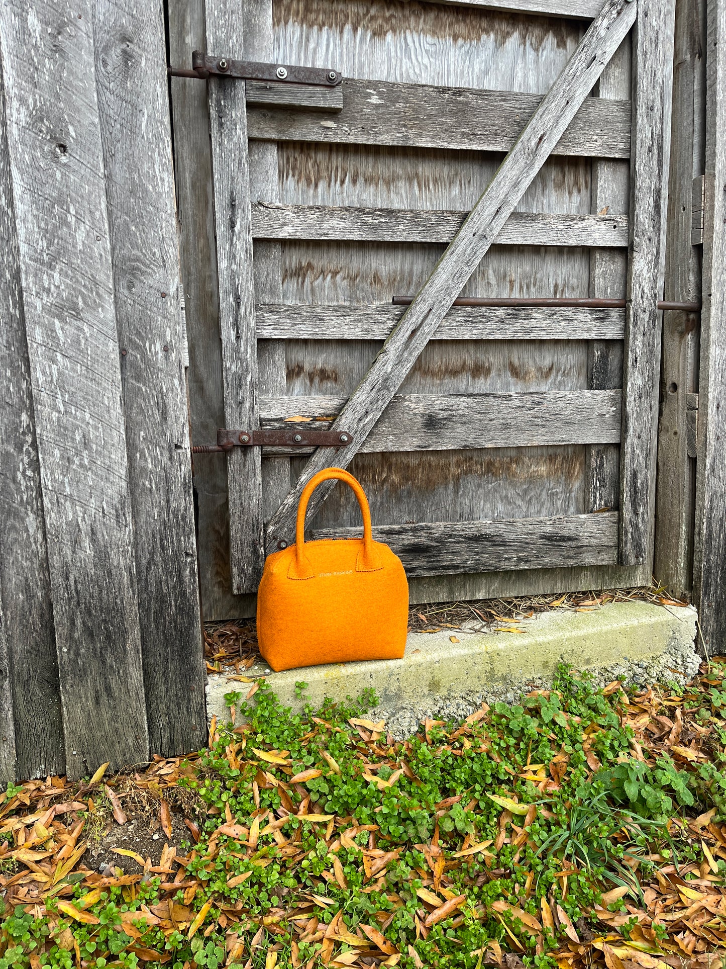 An orange handbag rests against an old wooden gate, surrounded by green plants and fallen leaves.