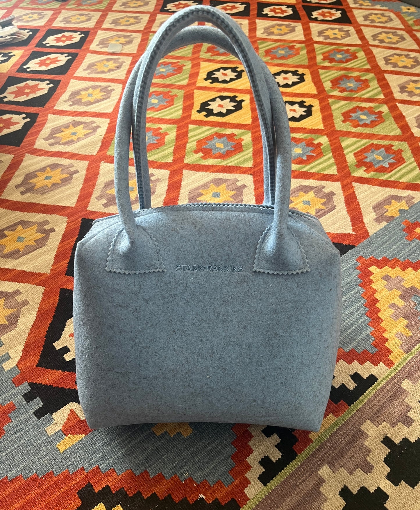 A light blue handbag with two handles placed on a colorful, patterned rug.