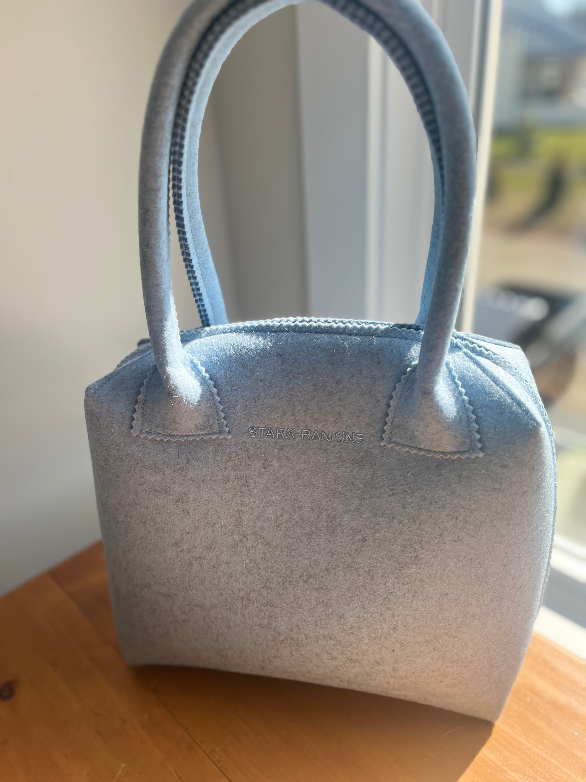 Light blue felt handbag with two handles, sitting on a wooden table by a window.