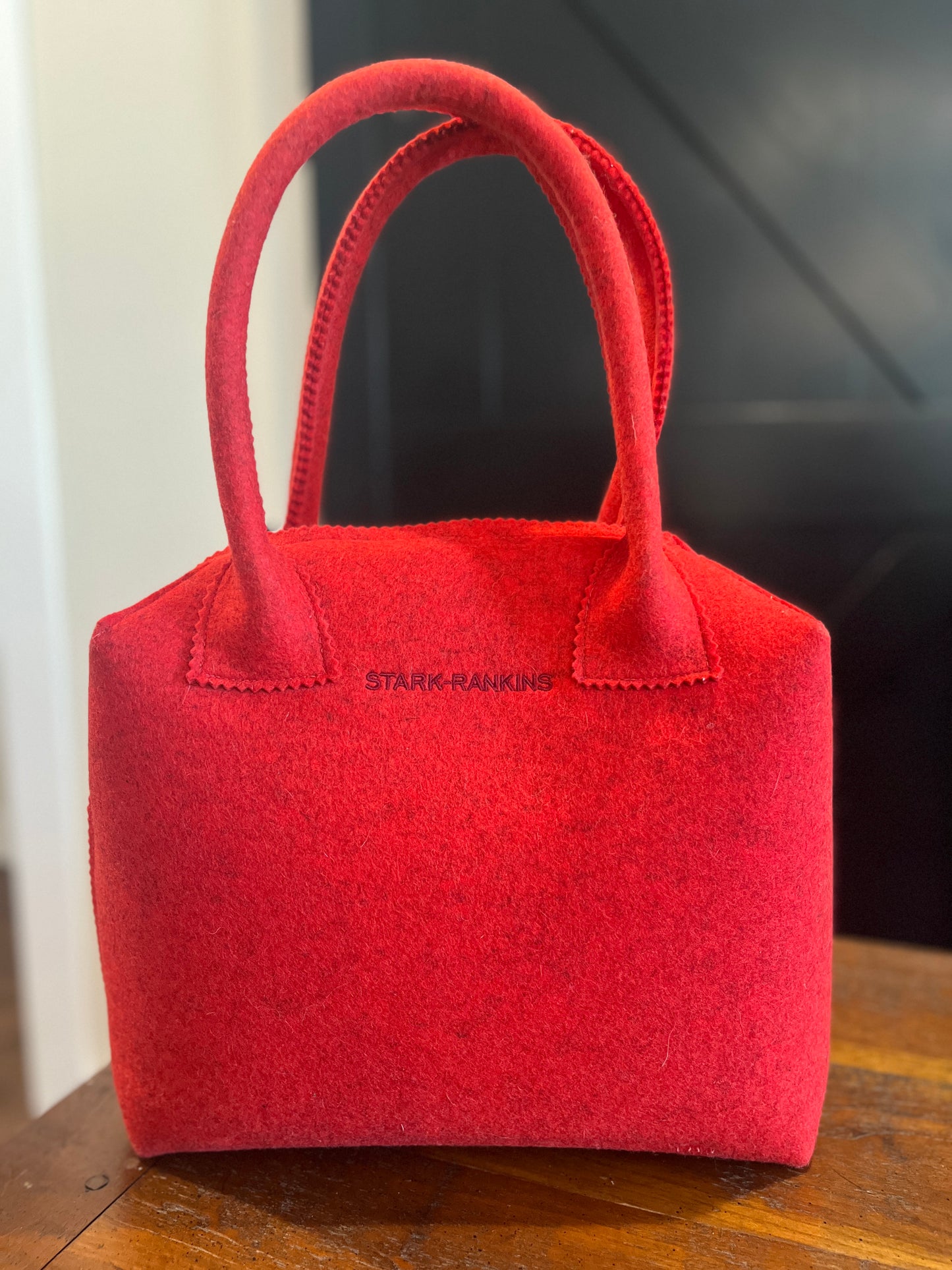 A red felt handbag with two handles, featuring the text "Stark-Rankins" on the side, placed on a wooden surface against a dark background.