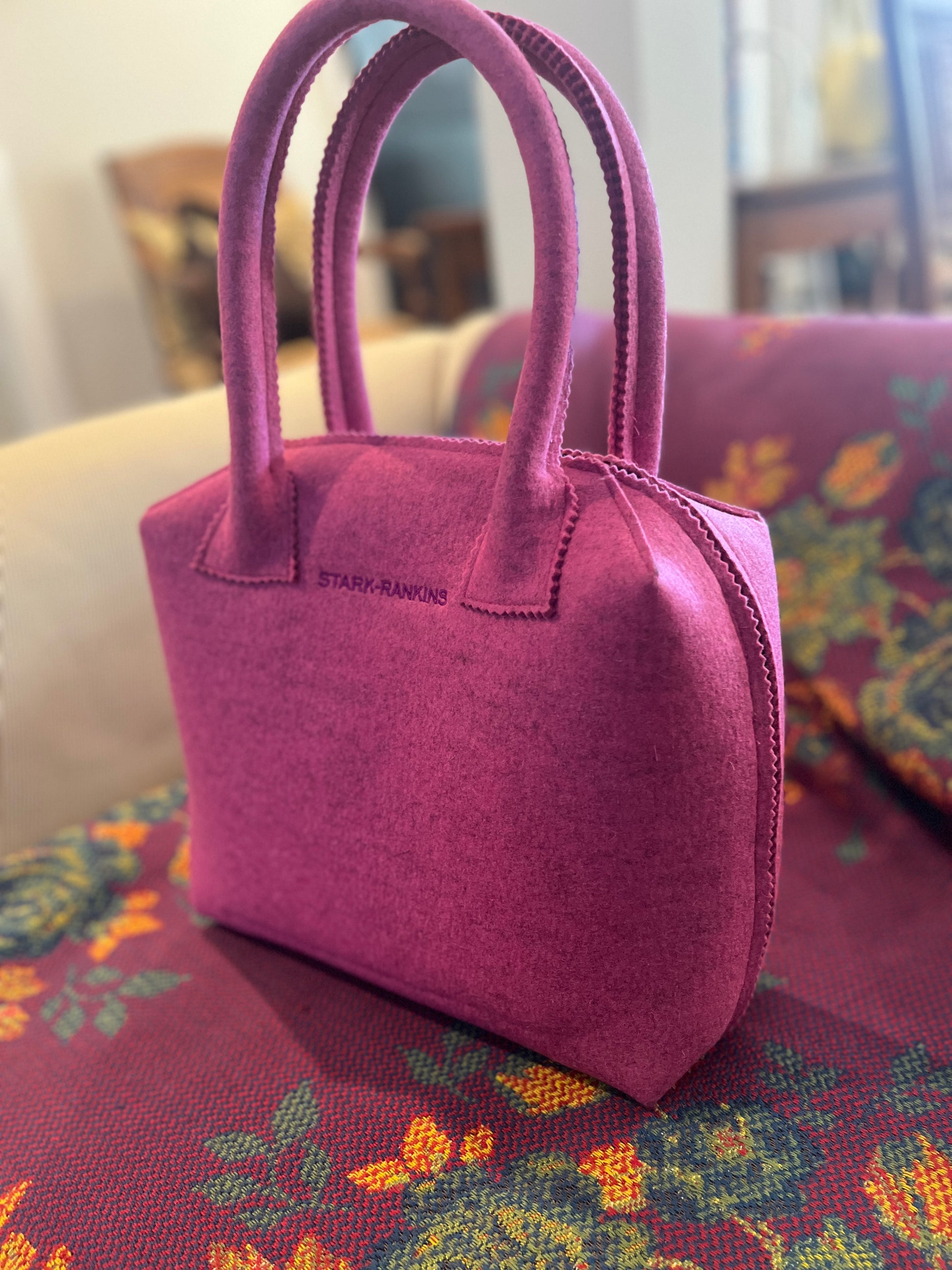 A magenta handbag with the label "Stark Rankins" rests on a floral-patterned fabric-covered surface.