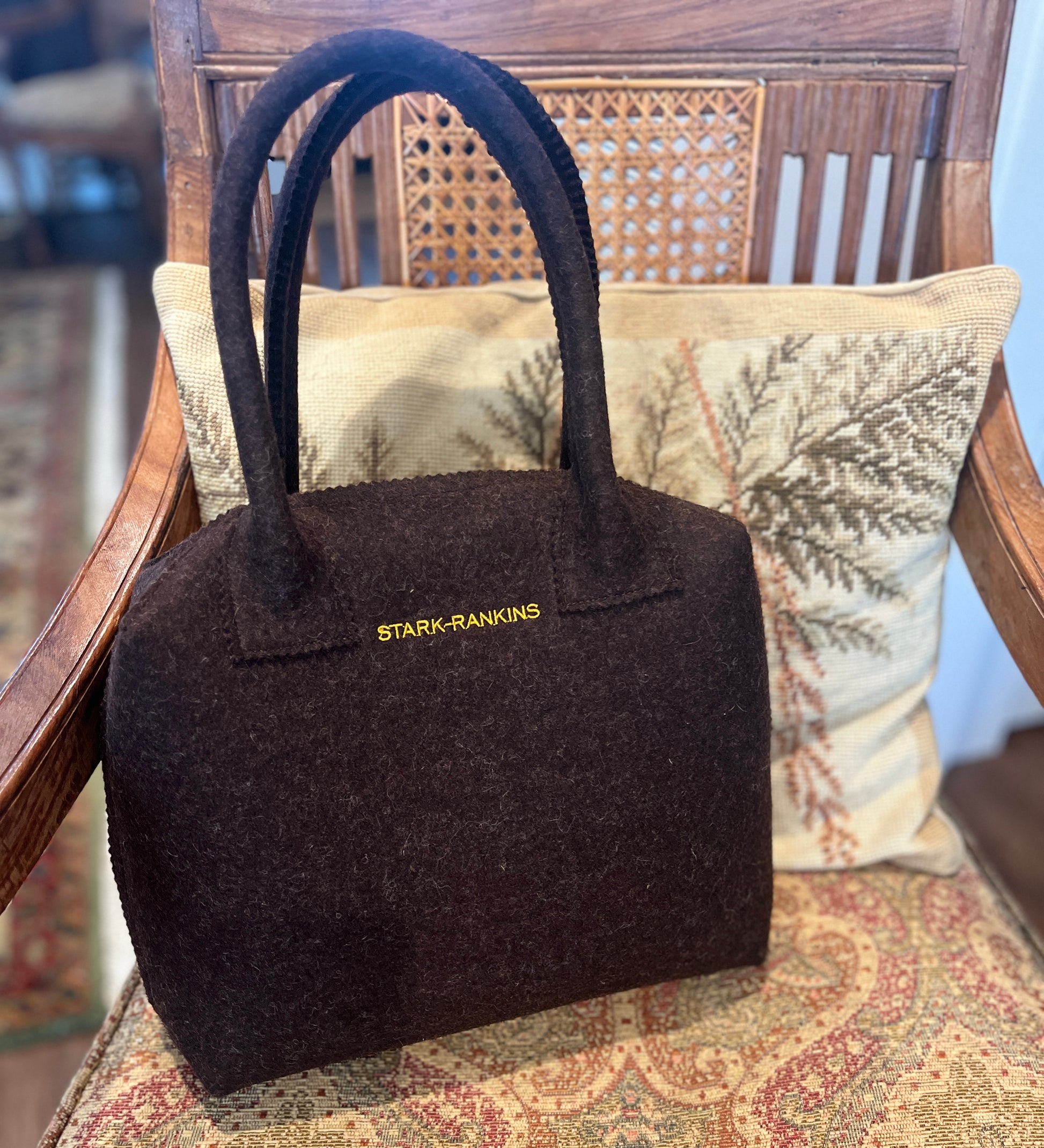 A brown felt handbag with "STARK-RANKINS" written on it is placed on a cushion on a wooden chair with wicker backrest.