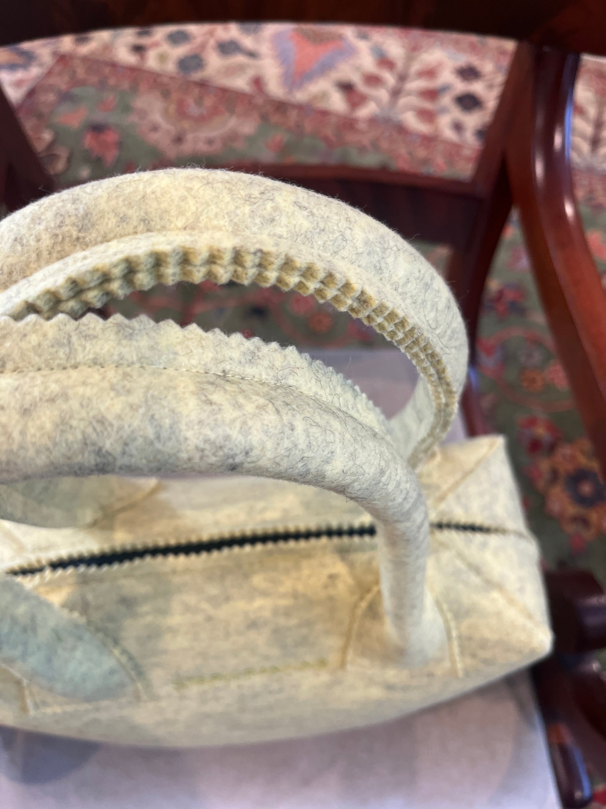 A close-up of a textured beige handbag with handles, positioned on a wooden chair. A patterned rug is partially visible in the background.