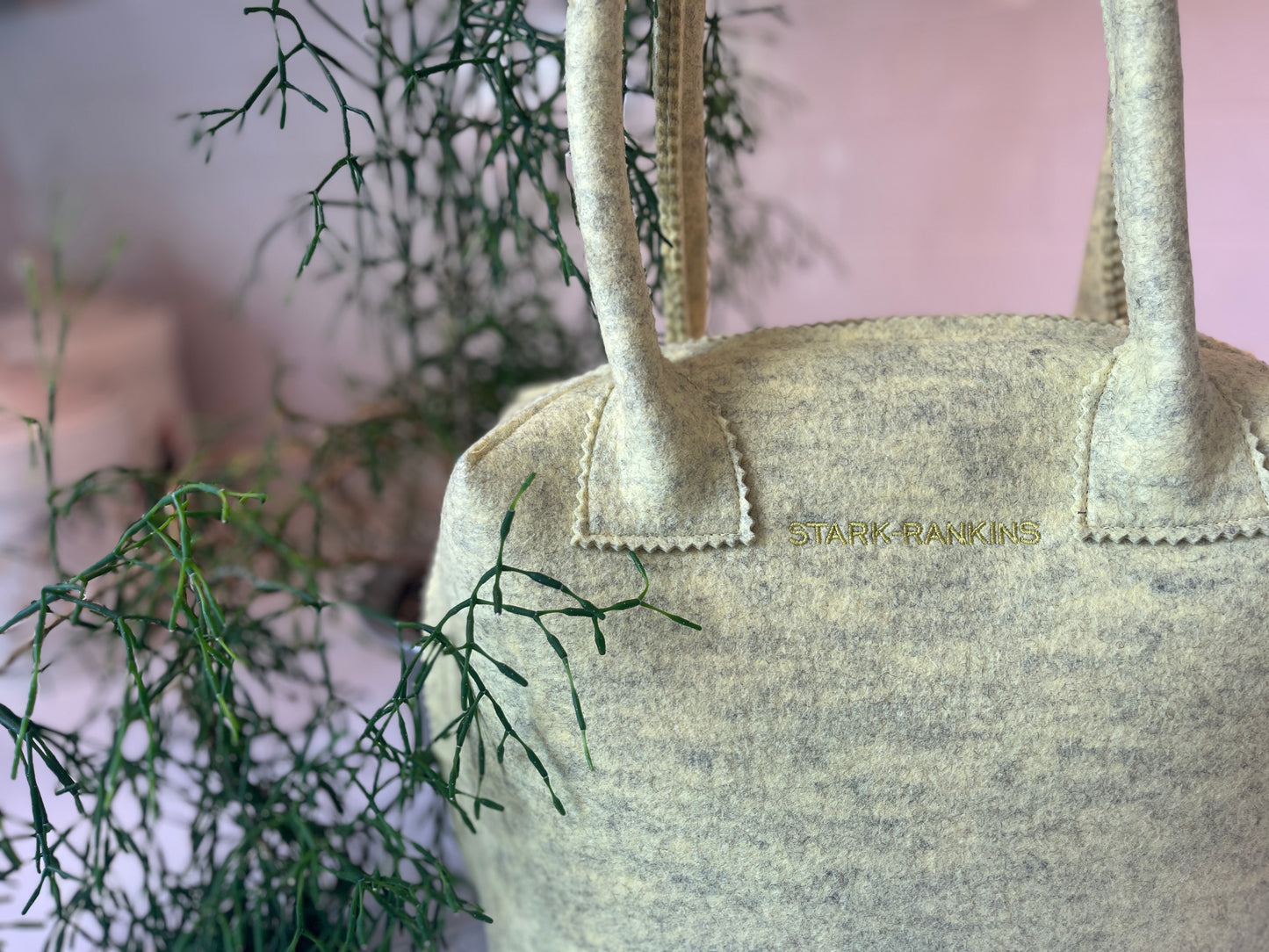 Cream-colored felt bag with handles, featuring "Stark Raving" embroidery, set against a backdrop of green branches.