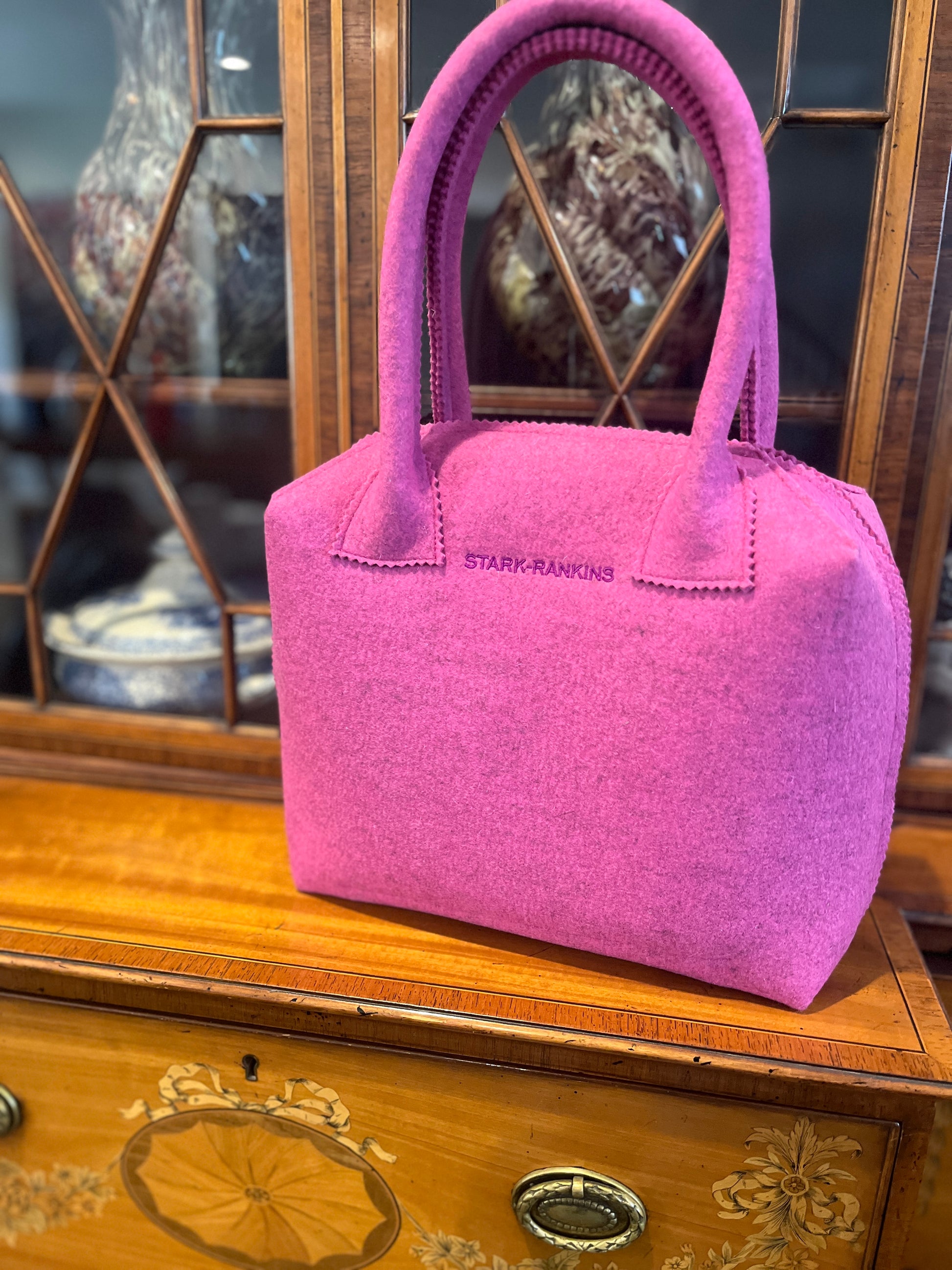 A pink felt handbag with the label "Stark-Rankins" sits on a wooden cabinet with decorative inlay, in front of a glass-front cabinet displaying vases.