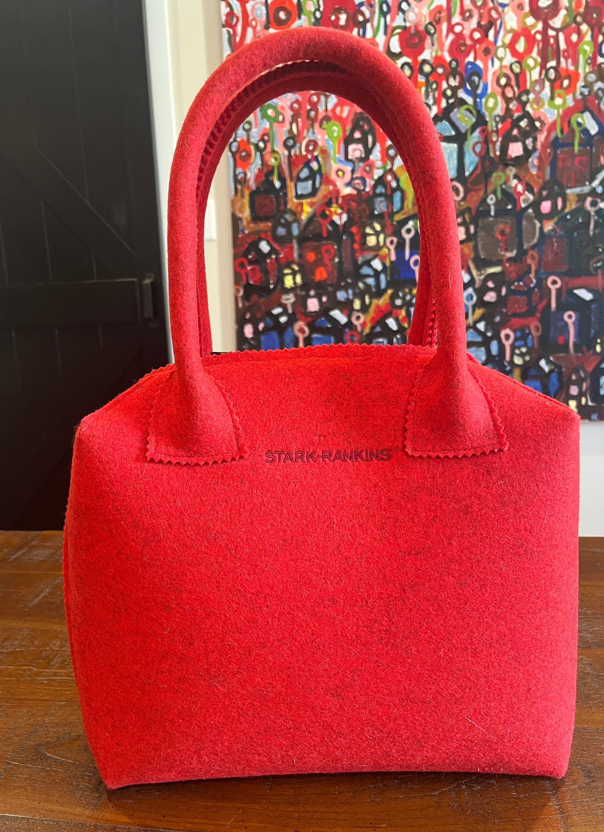 A red felt handbag on a wooden table, with a handle and subtle stitching detail. A colorful abstract painting is in the background.