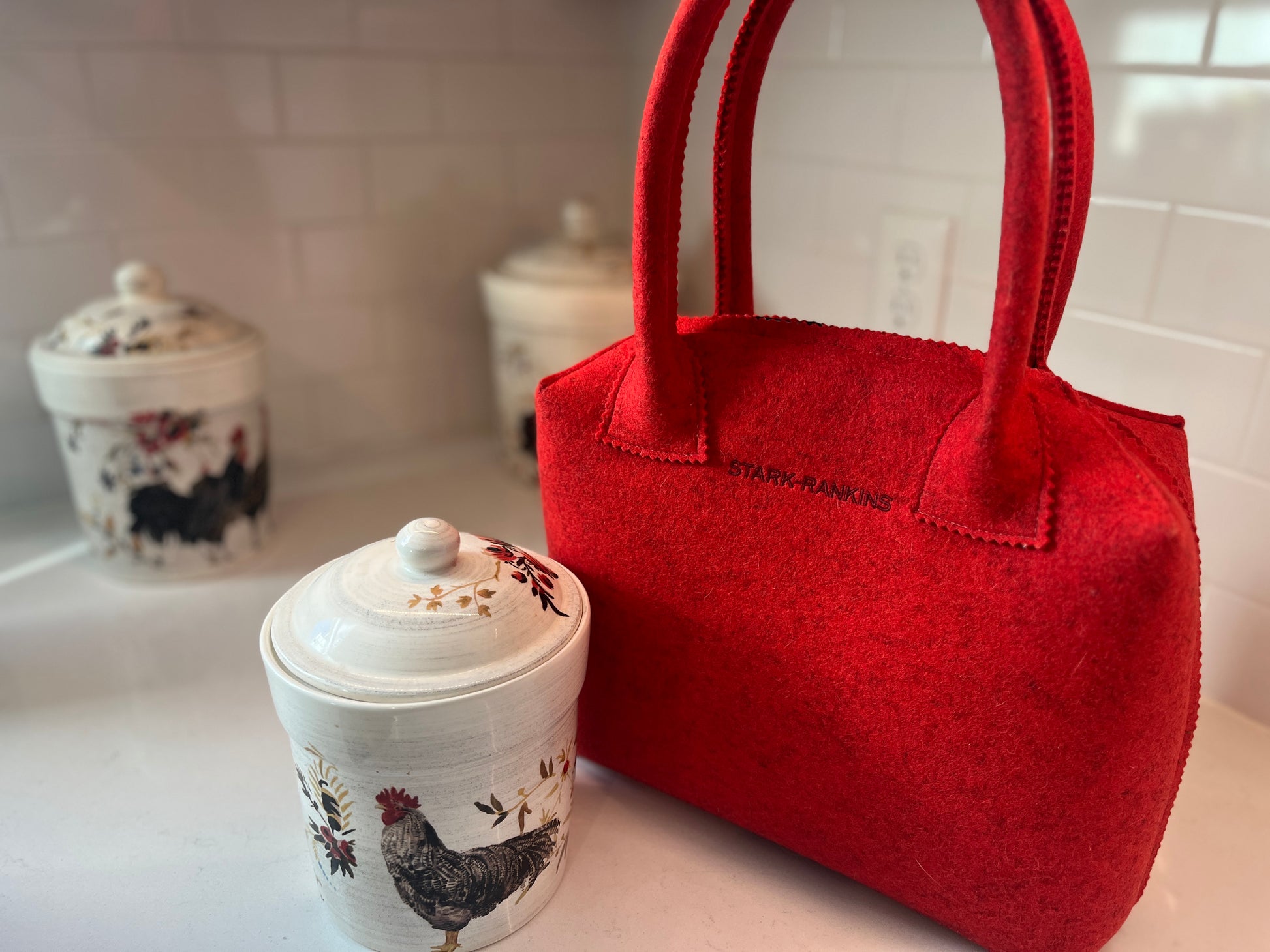 A red felt bag sits on a kitchen counter next to two ceramic containers adorned with rooster designs.