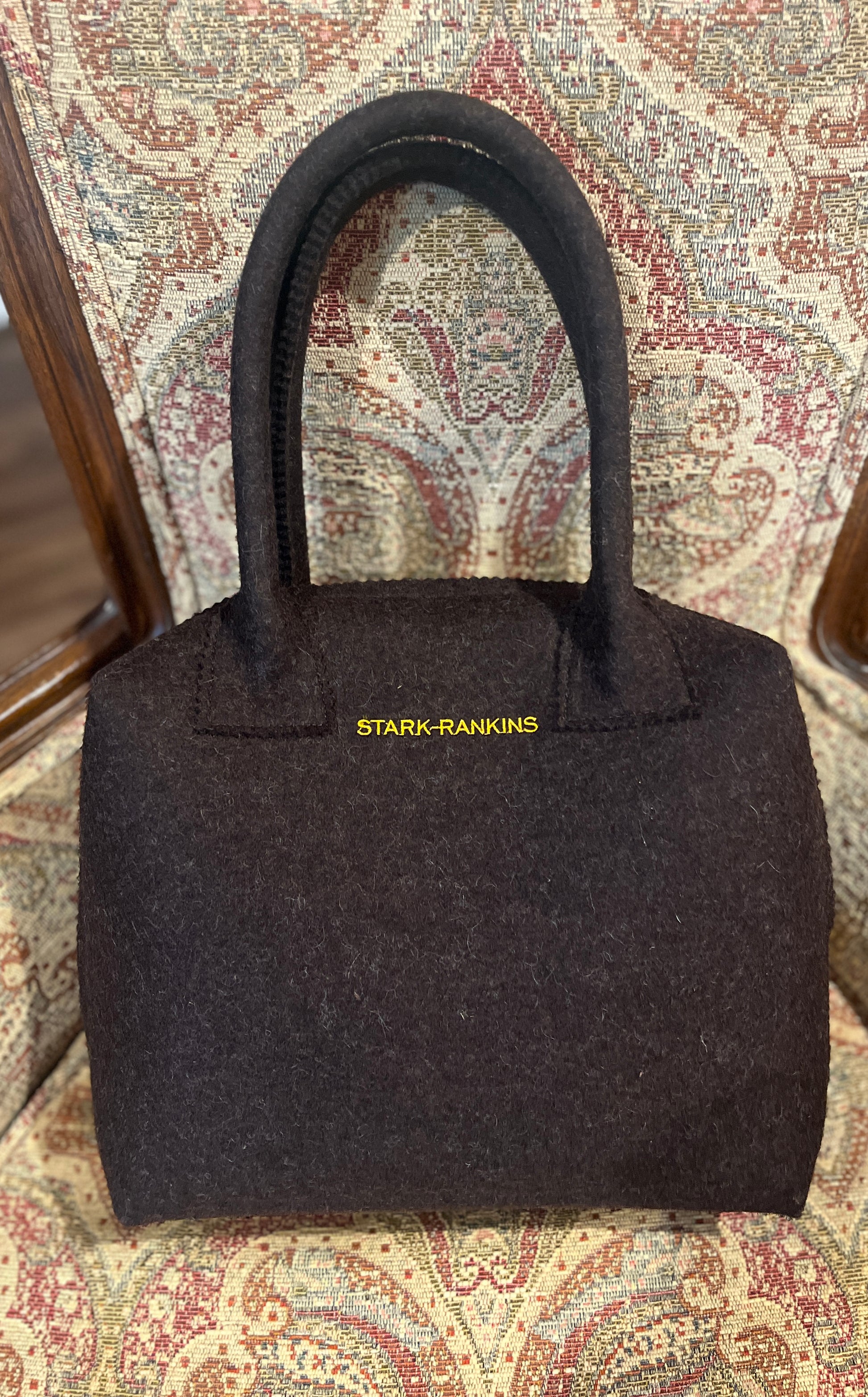 A black handbag with "STARK-RANKINS" in yellow text sits on a chair with a patterned fabric.