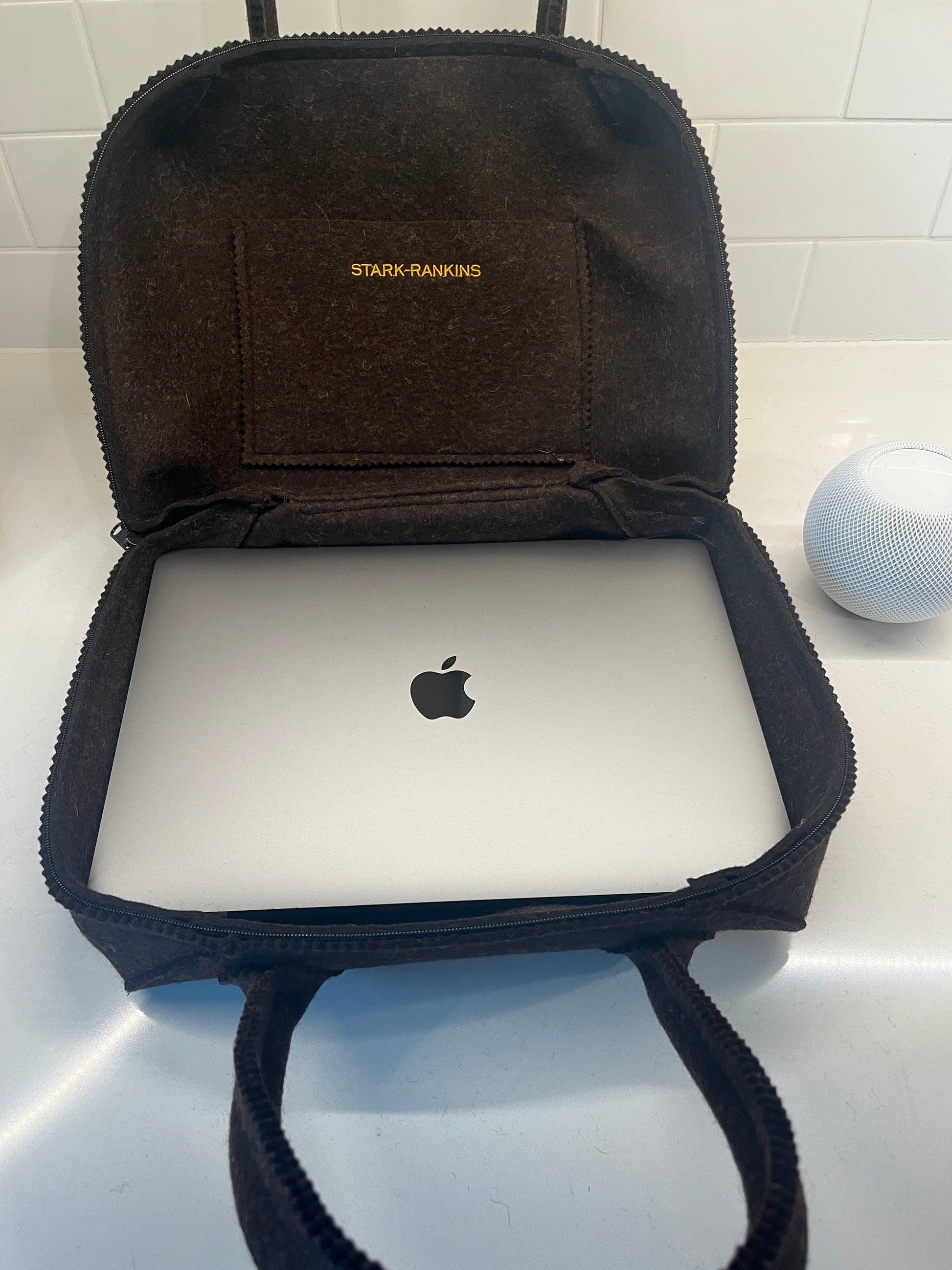 A laptop with an Apple logo inside an open brown felt bag on a white surface. A small speaker is visible in the background.