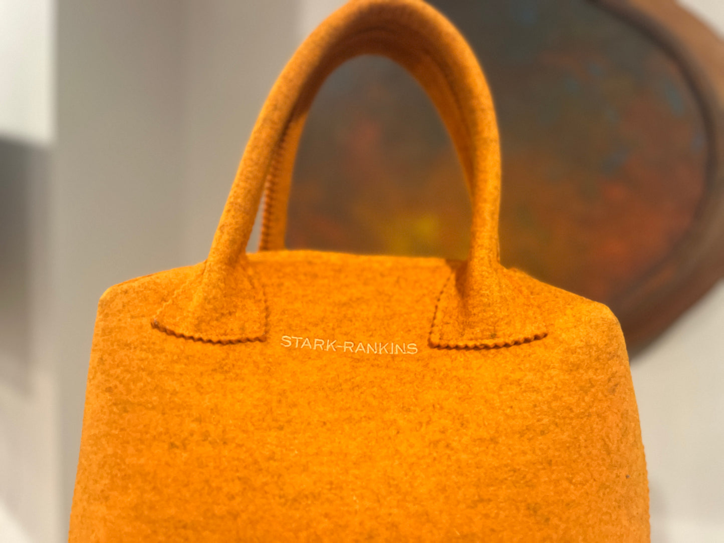 A bright orange felt handbag with sturdy handles, featuring the text "STARK-RANKINS" in small letters. A blurred, abstract background is visible behind it.