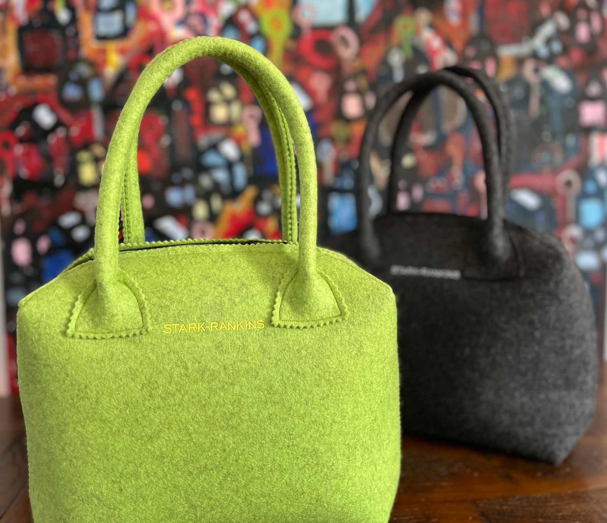 Two felt handbags on a wooden surface; one green and one black. The green bag has "Stark-Rankins" written on it. A colorful, abstract painting is in the background.