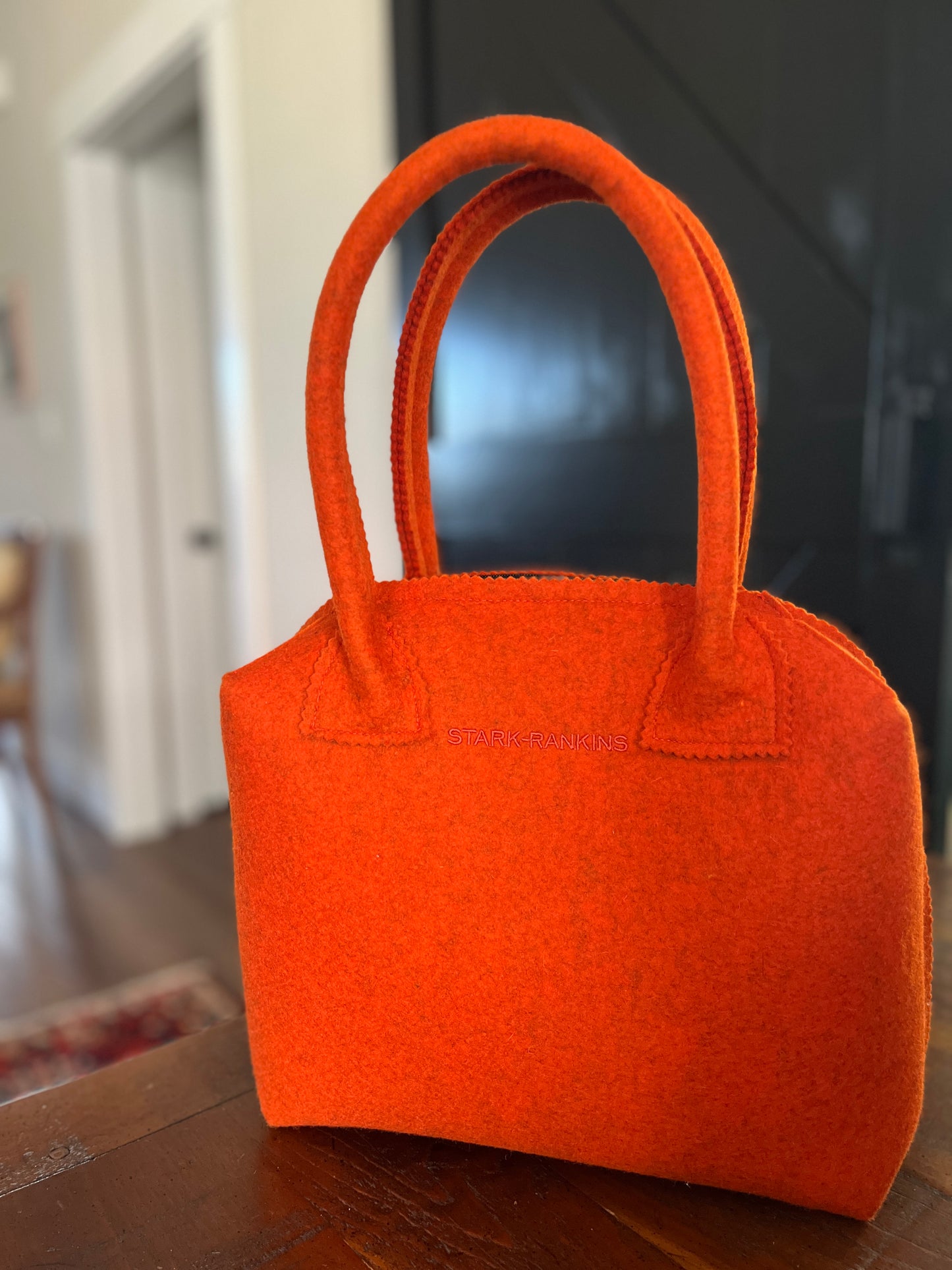 An orange felt handbag with two handles sits on a wooden surface in a room with blurred background elements.