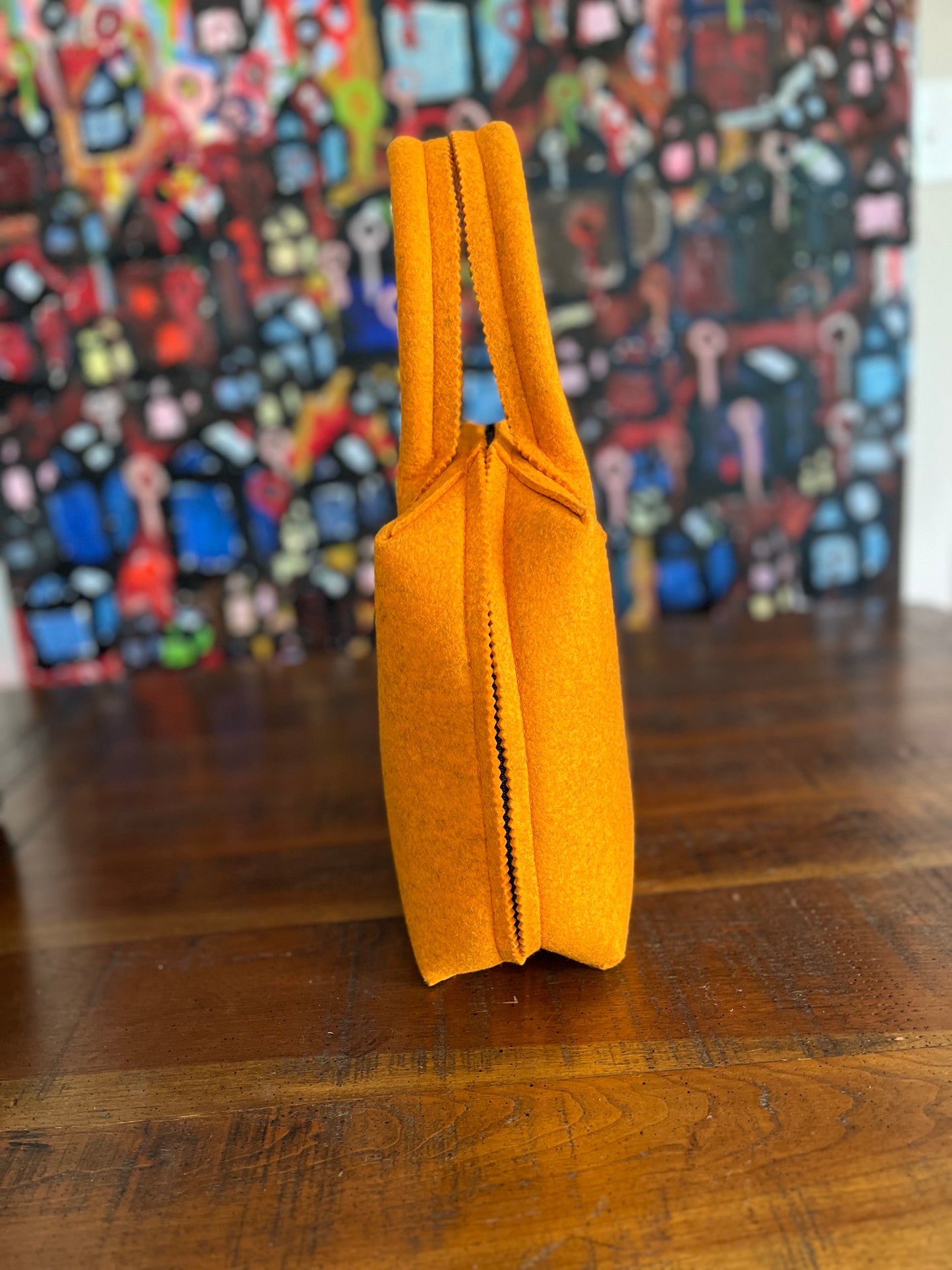 Side view of an orange felt handbag with two straps, placed on a wooden surface, against a colorful abstract background.