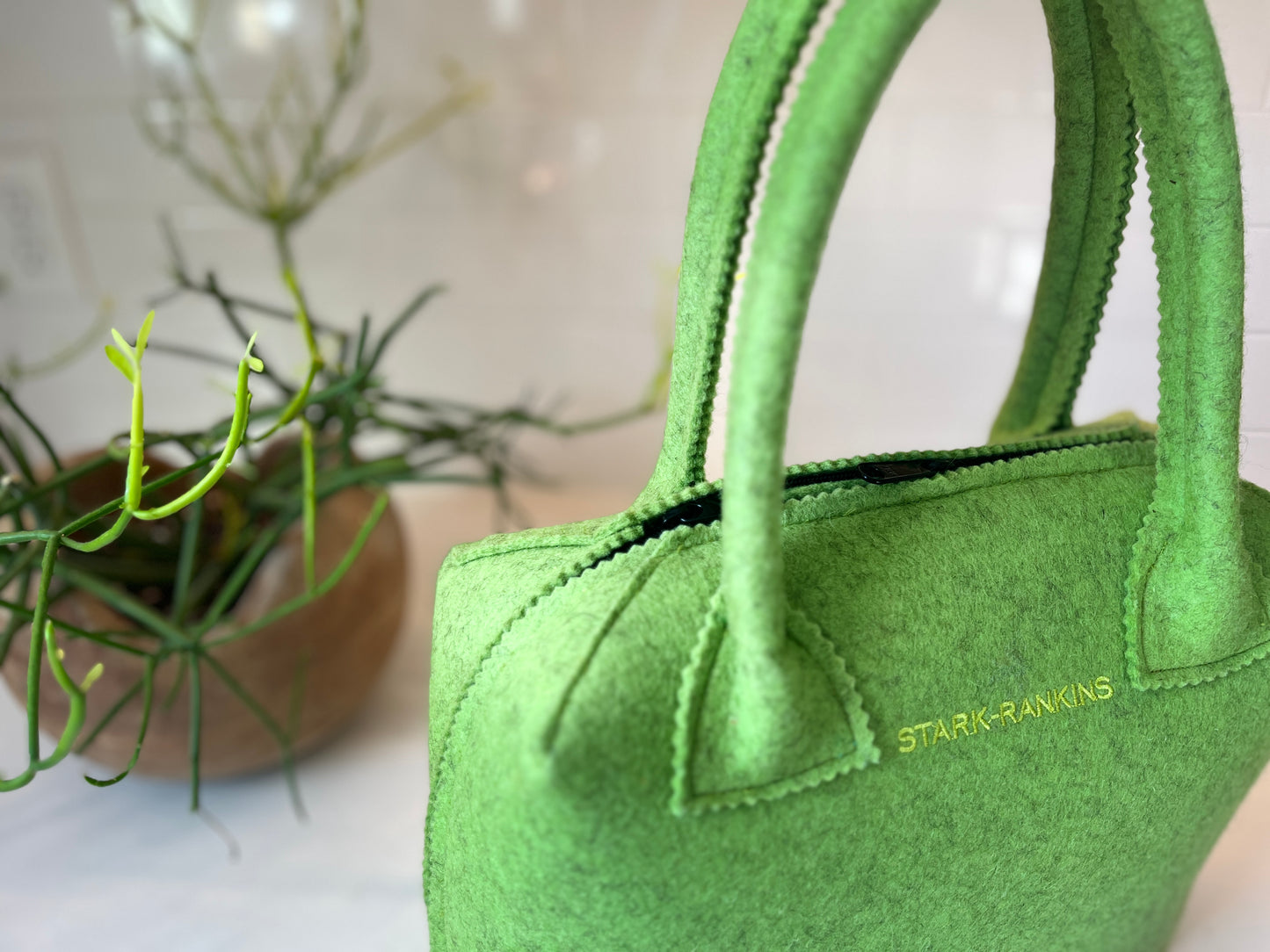 A green felt handbag with the name "Stark+Rankins" against a white tiled background and a plant in a wooden bowl.