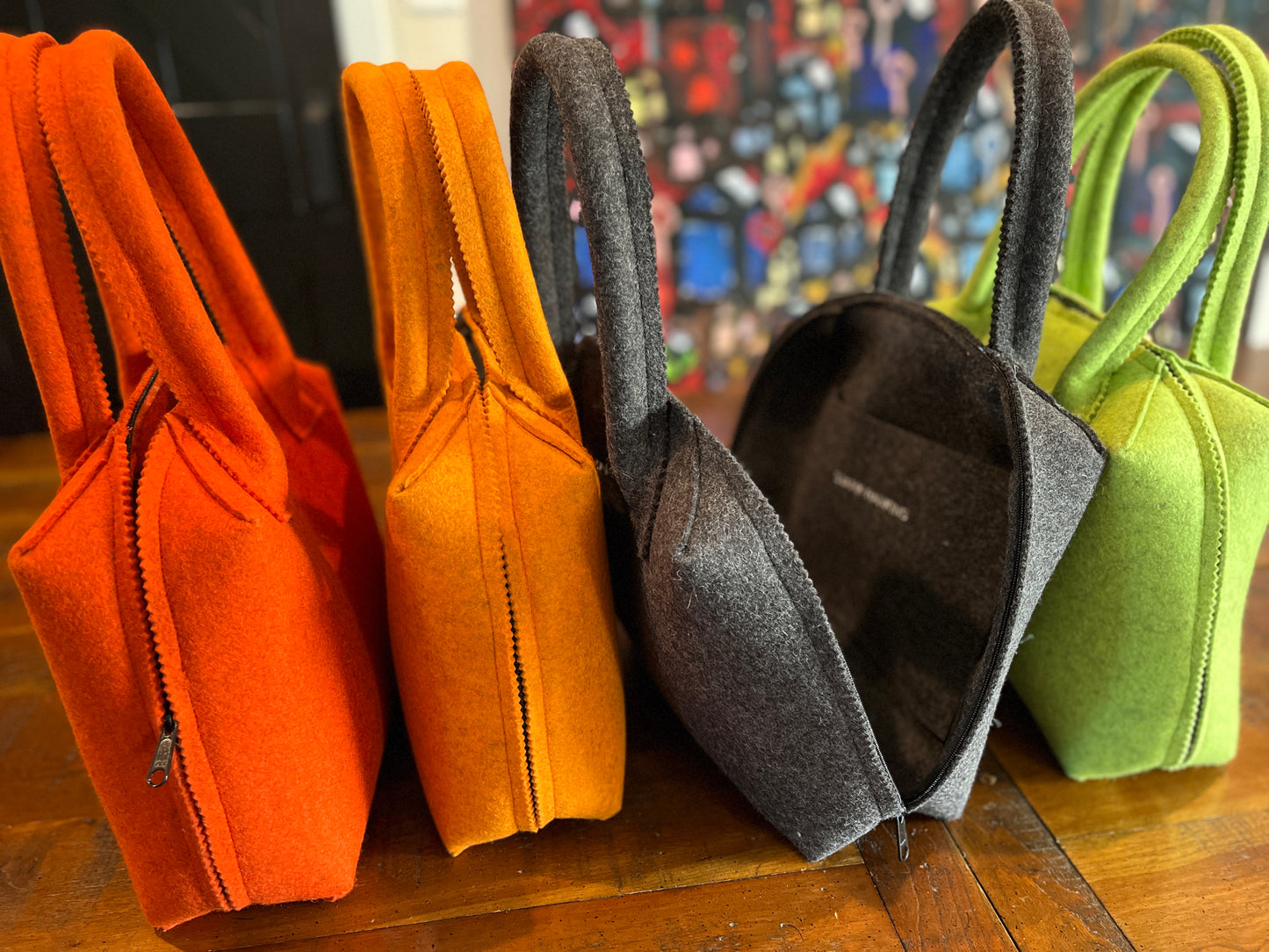 Four felt handbags, in orange, yellow, gray, and green, stand upright on a wooden table.