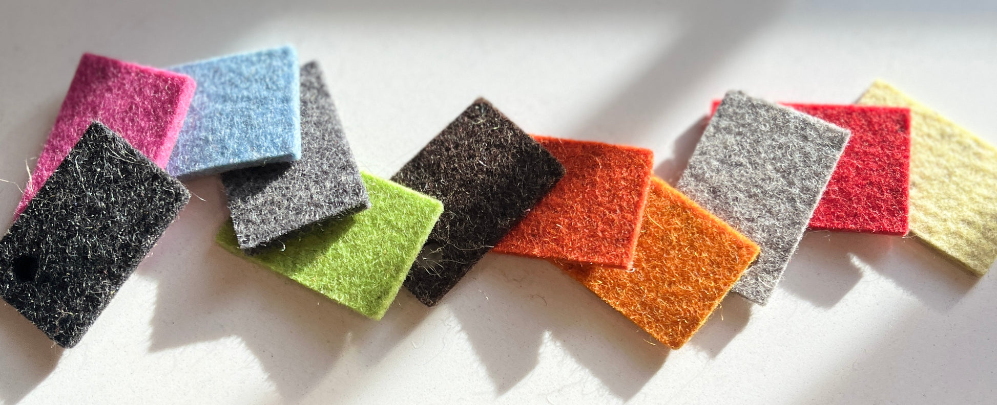 A row of 12 small felt squares in various colors, including blue, pink, gray, green, red, orange, and yellow, arranged on a white surface.